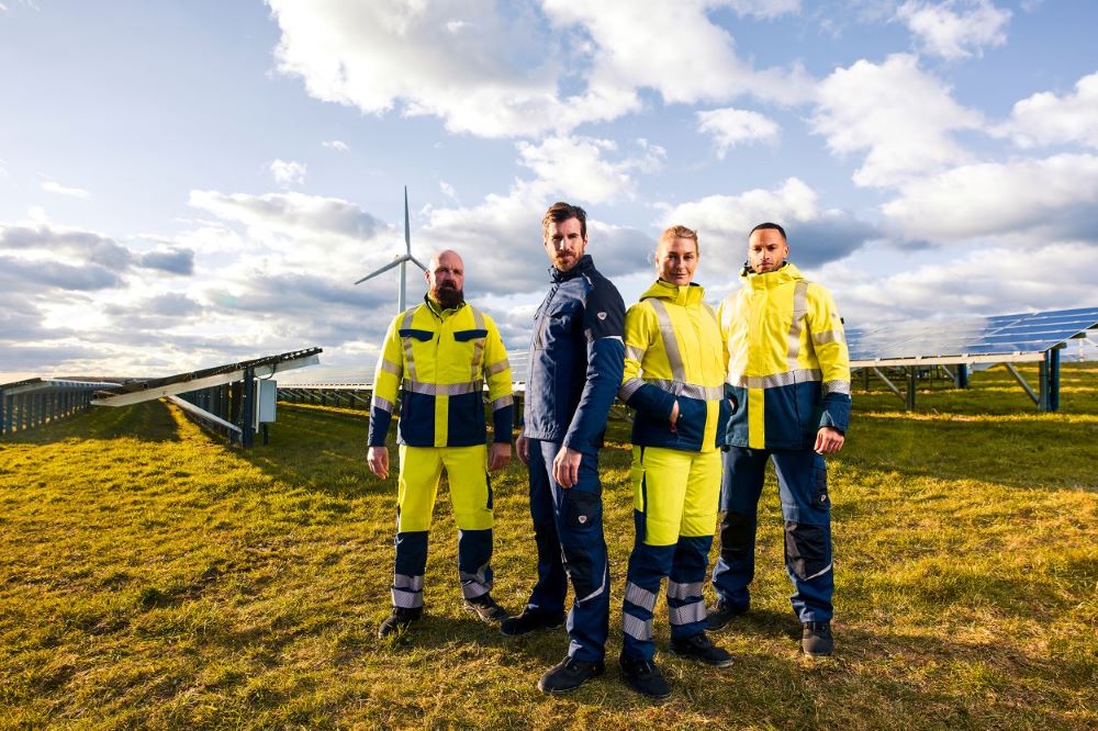 Quatre personnes portant des vêtements de travail modernes à haute visibilité devant des installations d'énergie solaire et éolienne.